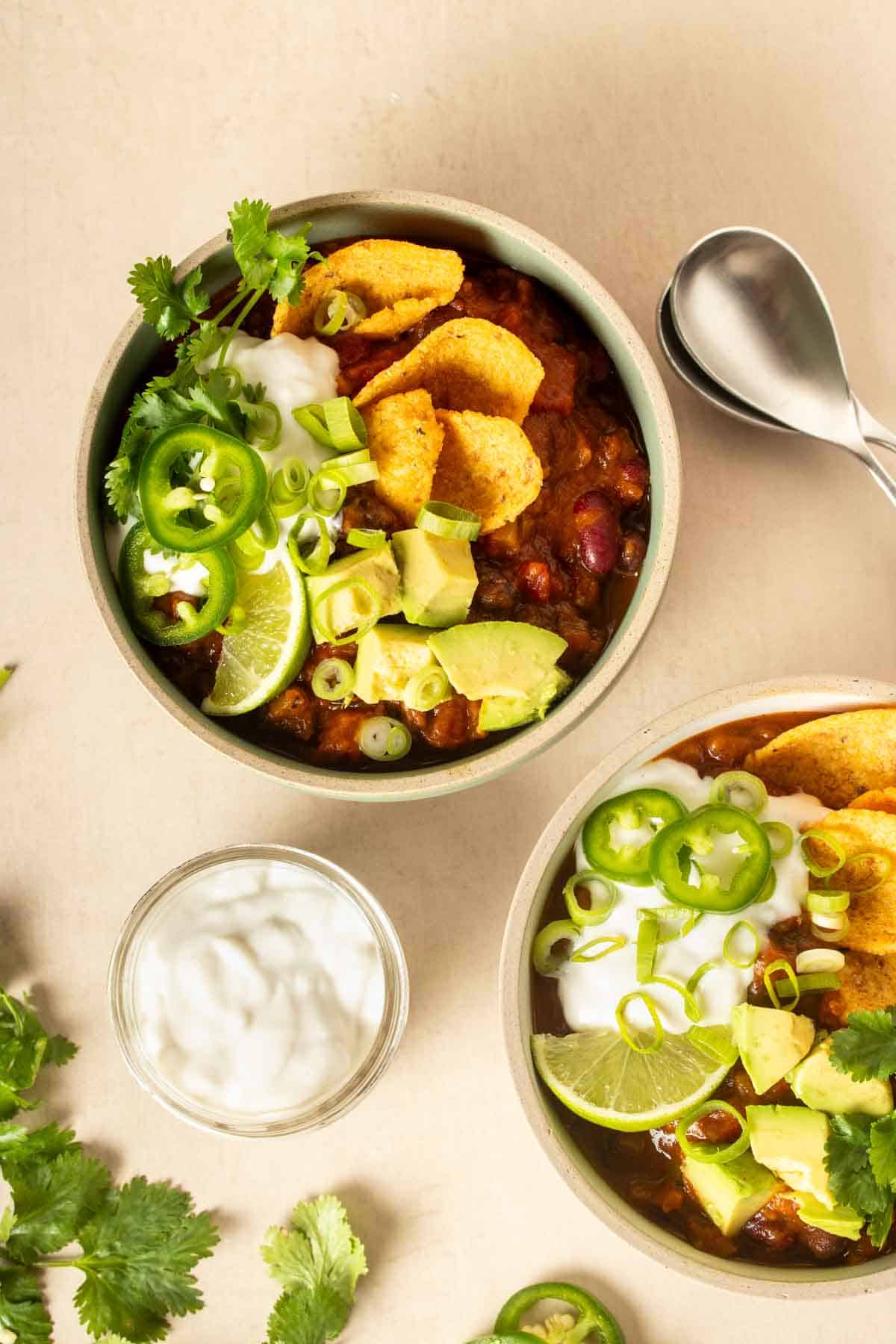 Two bowls of chili topped with ingredients like corn chips and avocado with spoons and more ingredients next to them.
