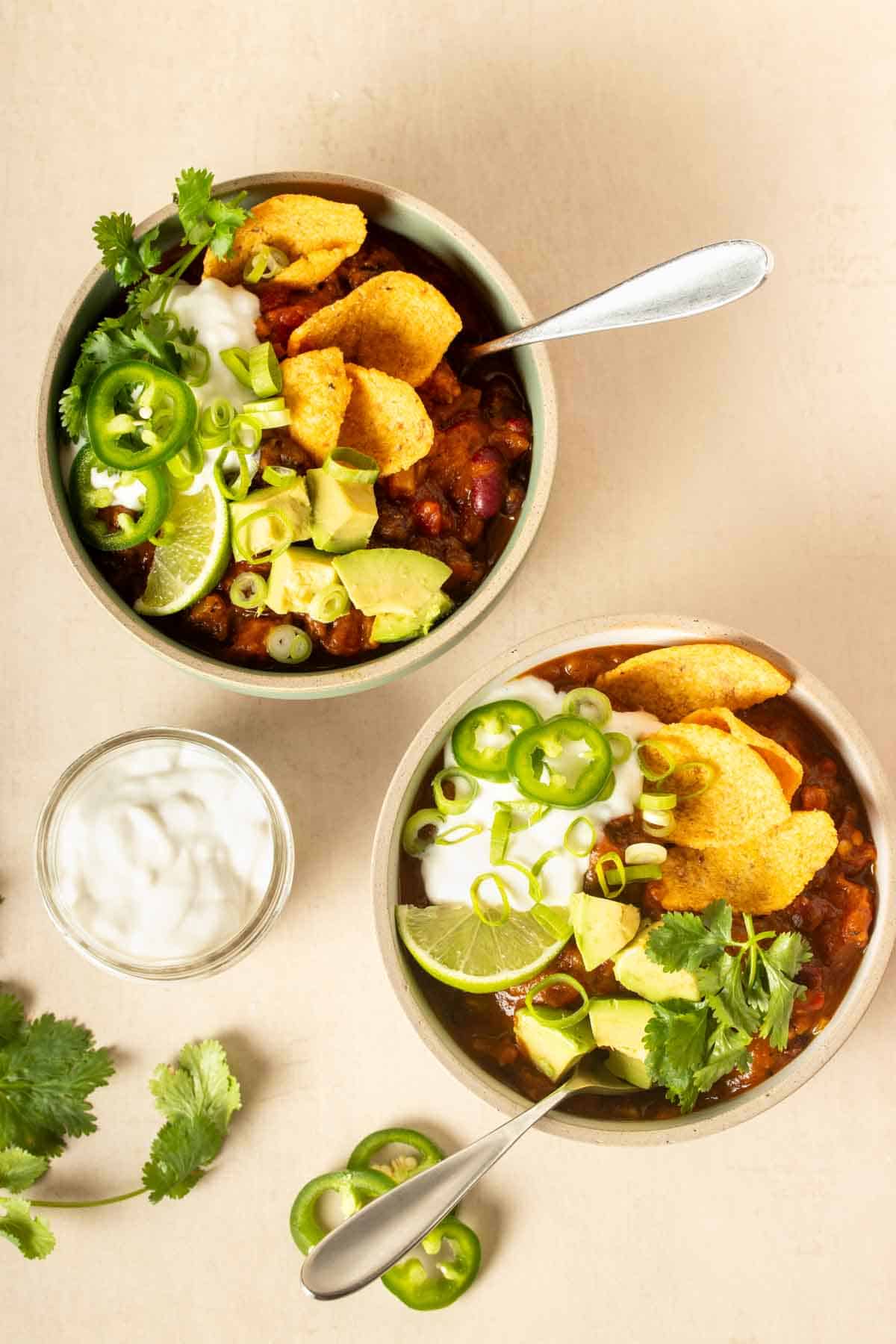 Top view of two bowls filled with chili and topped with corn chips, avocado, lime slice, jalapeno, sour cream and cilantro.