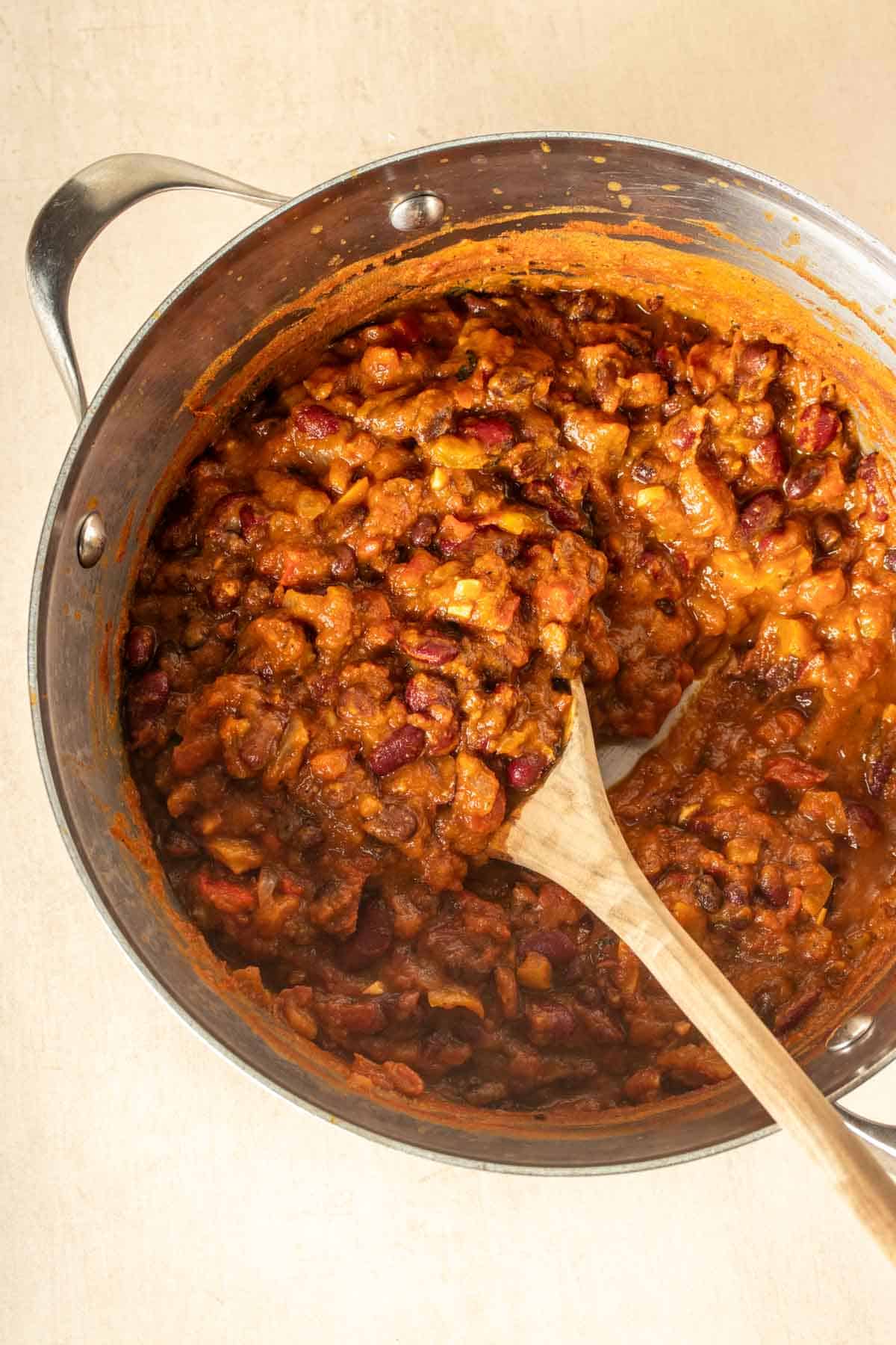 A wooden spoon mixing a pot of chili sitting on a tan surface.