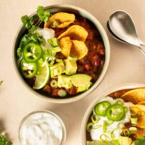 Chili in a bowl and topped with corn chips, avocado, sour cream, jalapeno and cilantro with a lime slice on it.