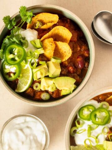 Chili in a bowl and topped with corn chips, avocado, sour cream, jalapeno and cilantro with a lime slice on it.