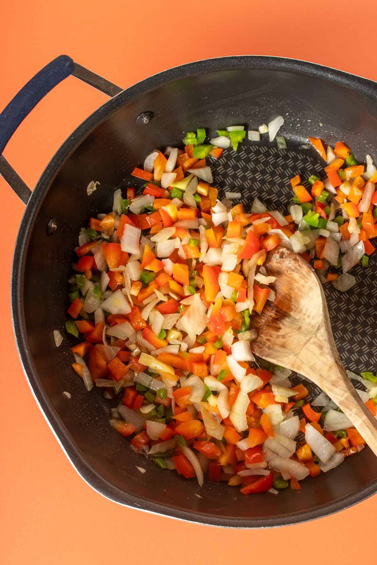 A pan with chopped peppers and onions being sauteed by a wooden spoon.