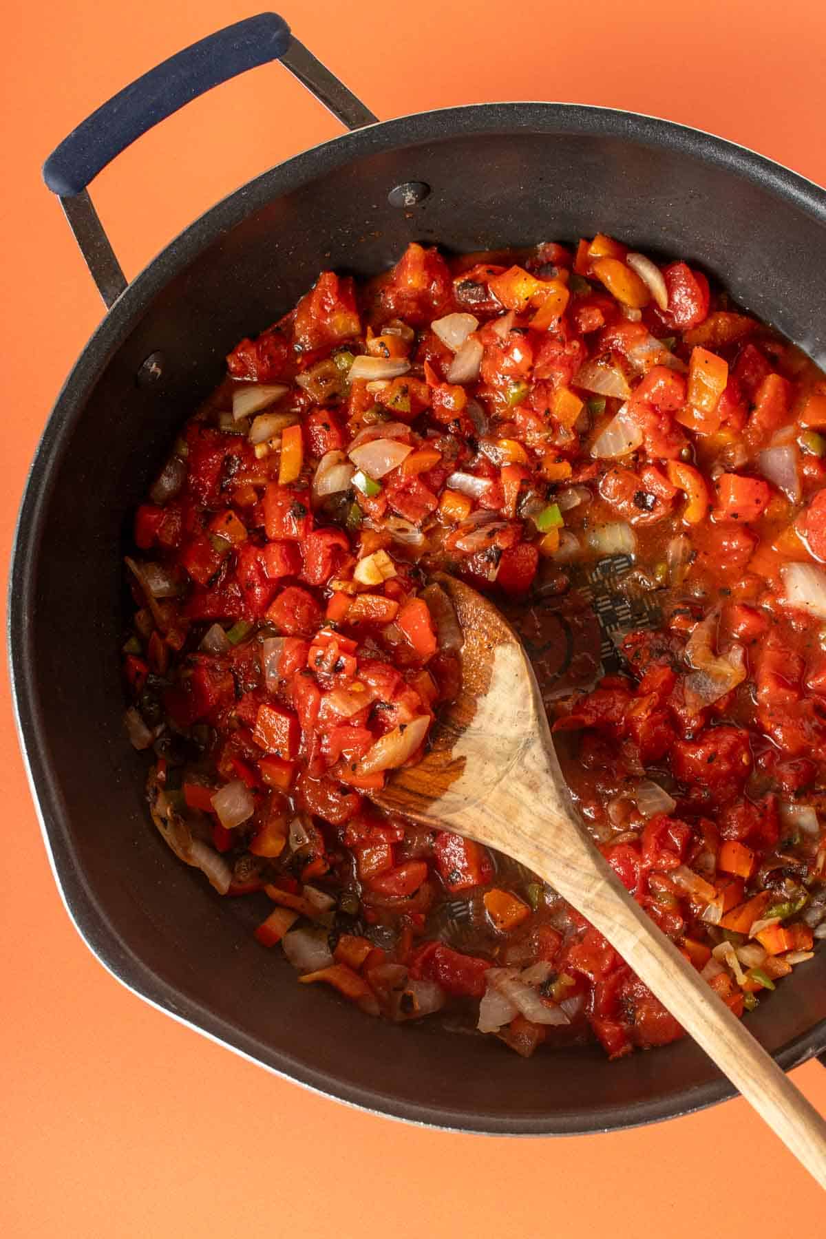 A wooden spoon mixing tomatoes, peppers and onions in a pan.