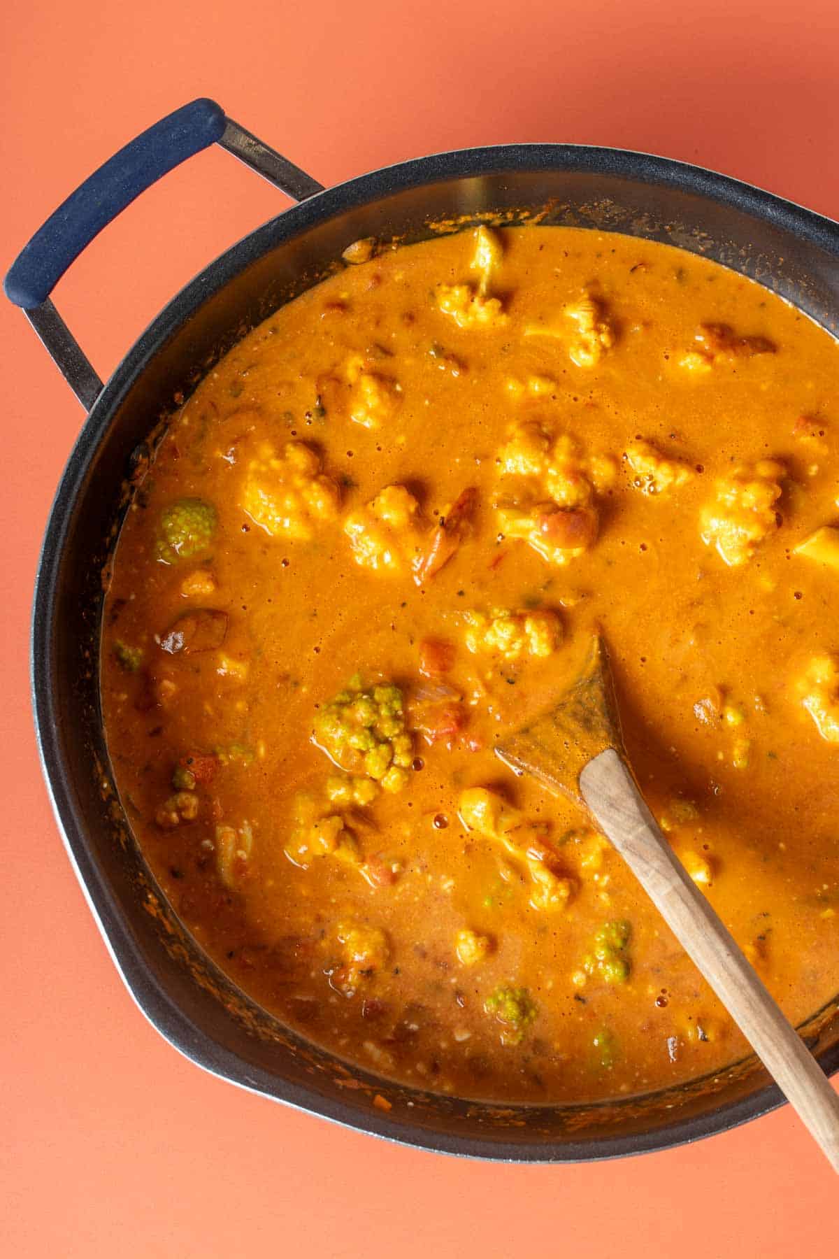 A wooden spoon mixing a red curry with chickpeas and cauliflower in a pan.