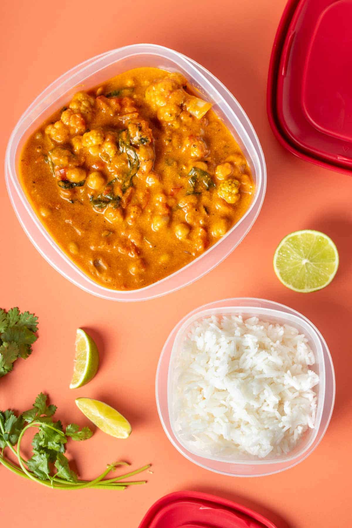 Square plastic containers filled with white rice and curry and red lids next to them with lime slices and cilantro.