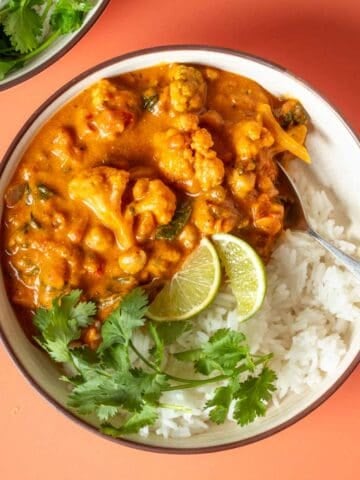 White rice topped with curry and cilantro sprigs and lime slices witting on an orange surface.