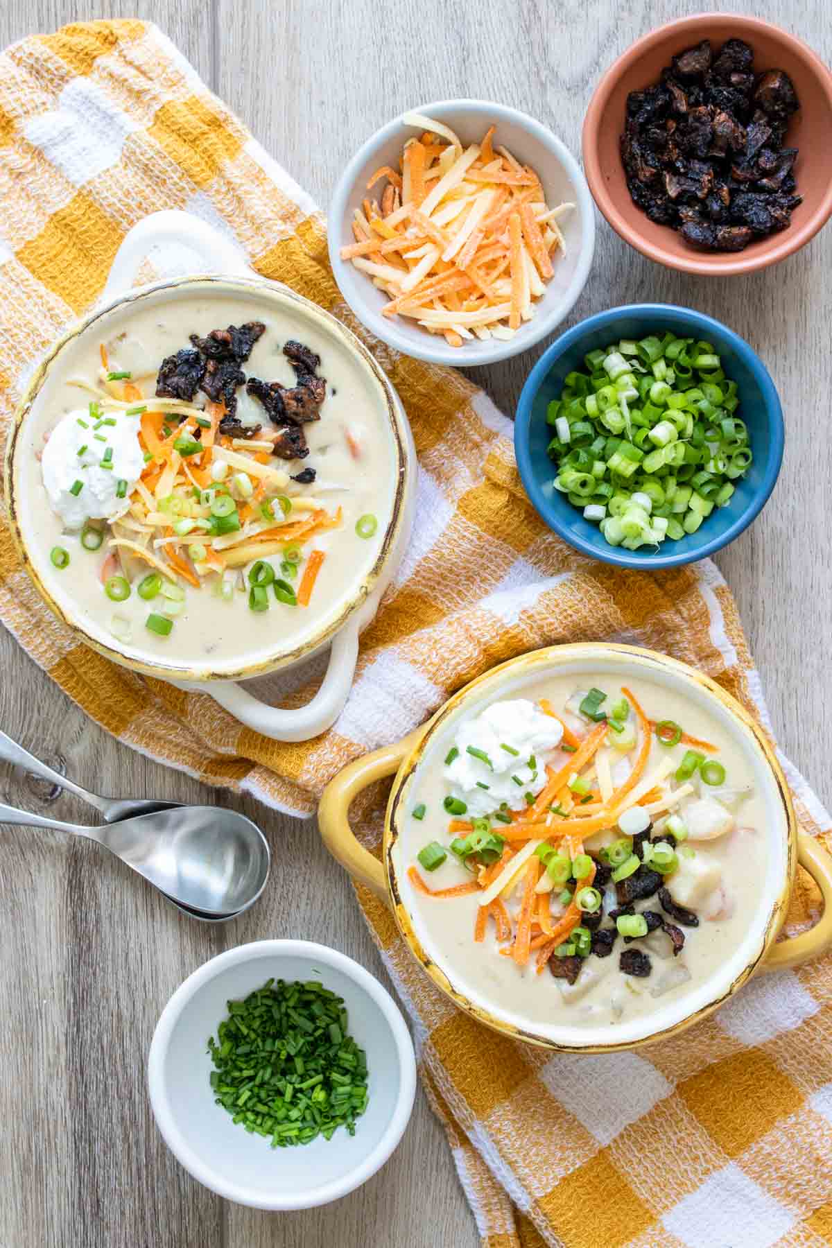 Two bowls of fully loaded potato soup on a yellow and white checkered towel next to toppings.