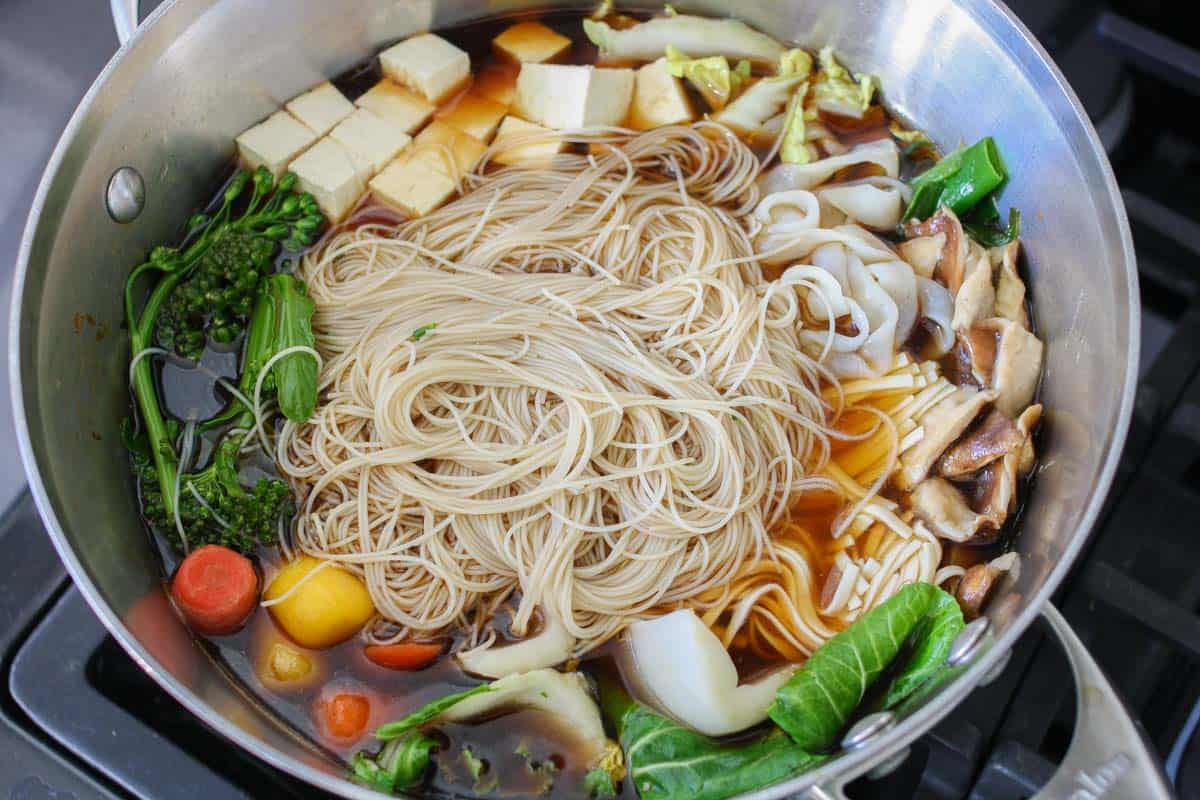 A metal pot with a broth that has noodles in the center and veggies surrounding them.