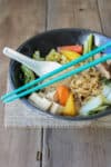 Turquoise chopsticks resting on a black bowl with some veggie sukiyaki in it.