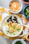 A photo of potato soup in a cream soup bowl on a yellow checked towel covered in toppings.