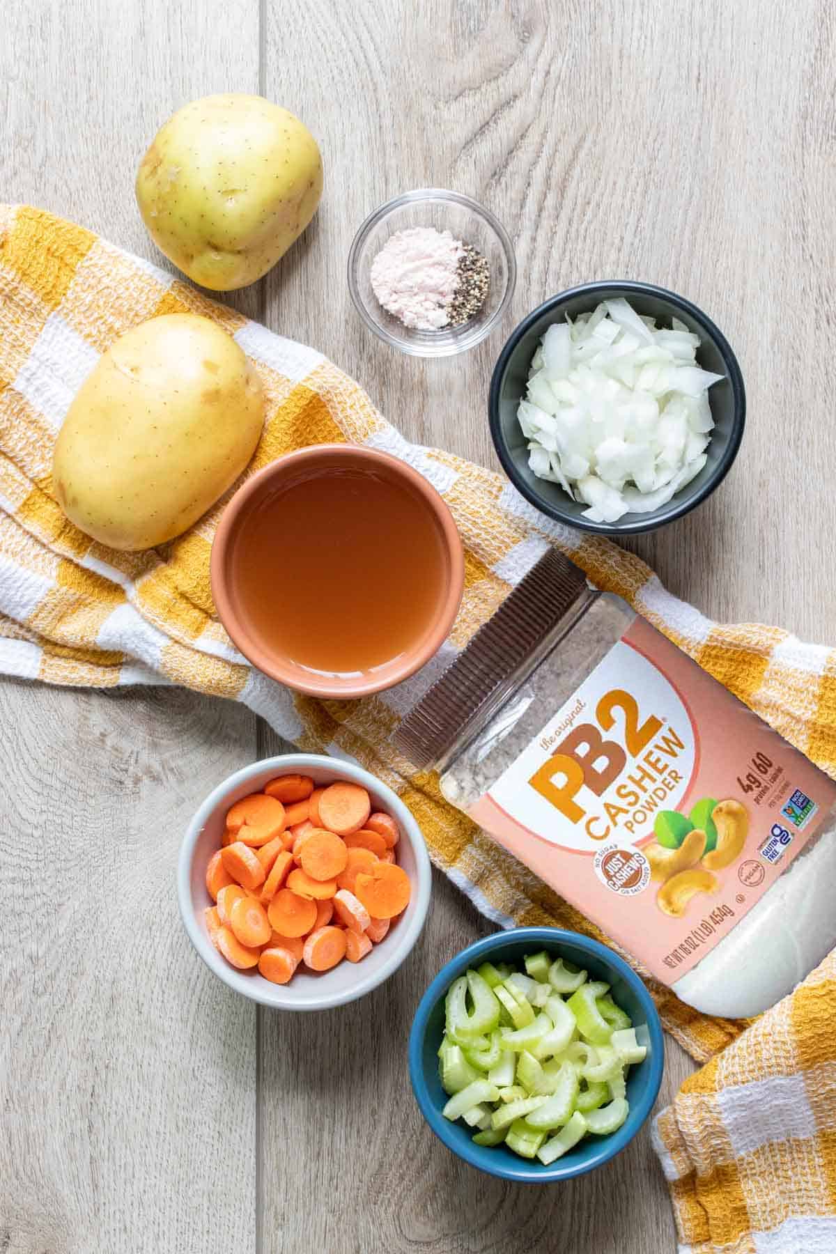 A container of PB2 cashew powder on a yellow and white towel with other ingredients needed to make a potato soup.