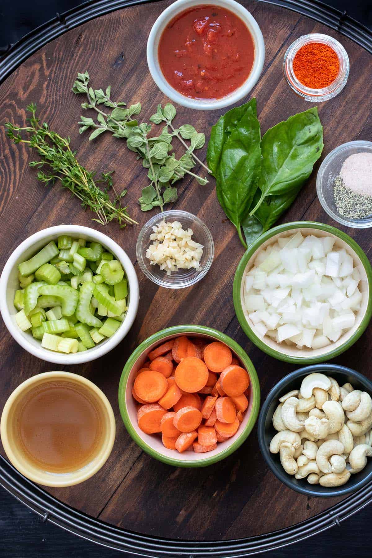 A wooden tray with colored bowls of ingredients for a cashew creamy based tomato soup with fresh herbs on it.