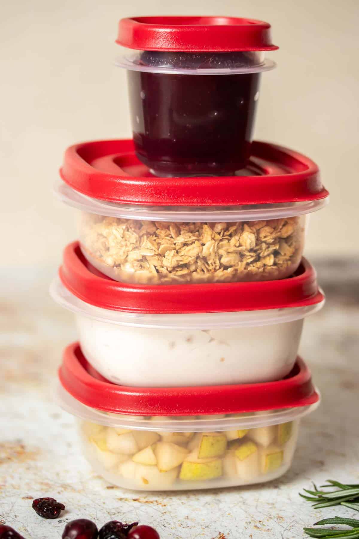 A pile of three large plastic containers with red tops and a smaller one on top, filled with fruit, yogurt, granola and jelly.