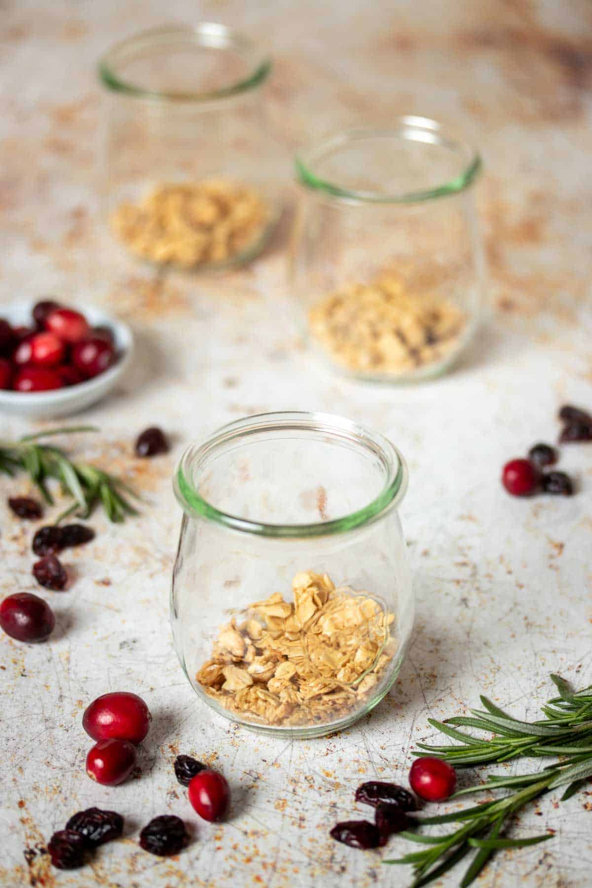 Granola at the bottom of three glass jars sitting on a tan speckled surface with cranberries and rosemary sprigs.