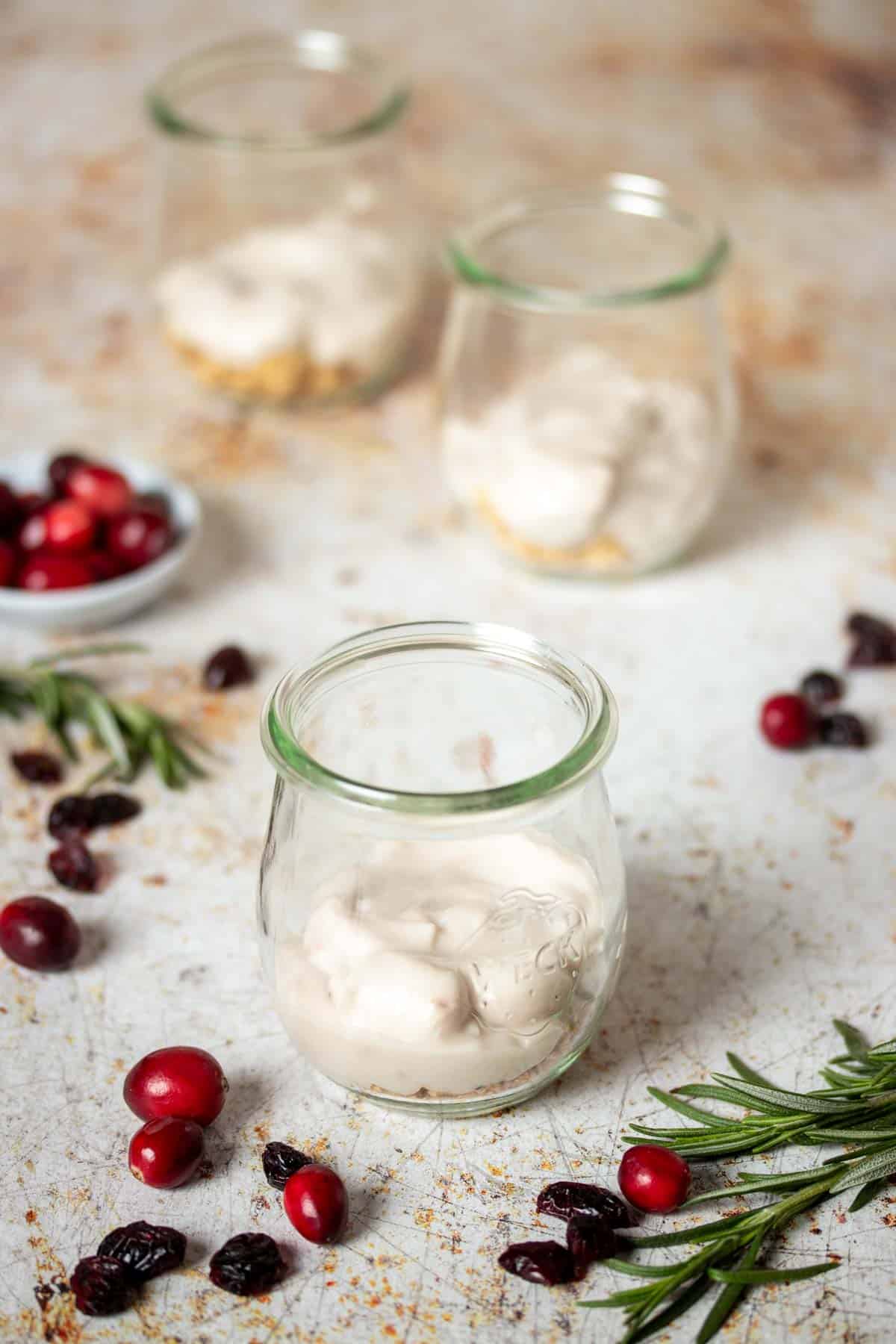 Breakfast parfait being layered with granola and yogurt in 3 glass jars with cranberries and rosemary sprigs next to them.