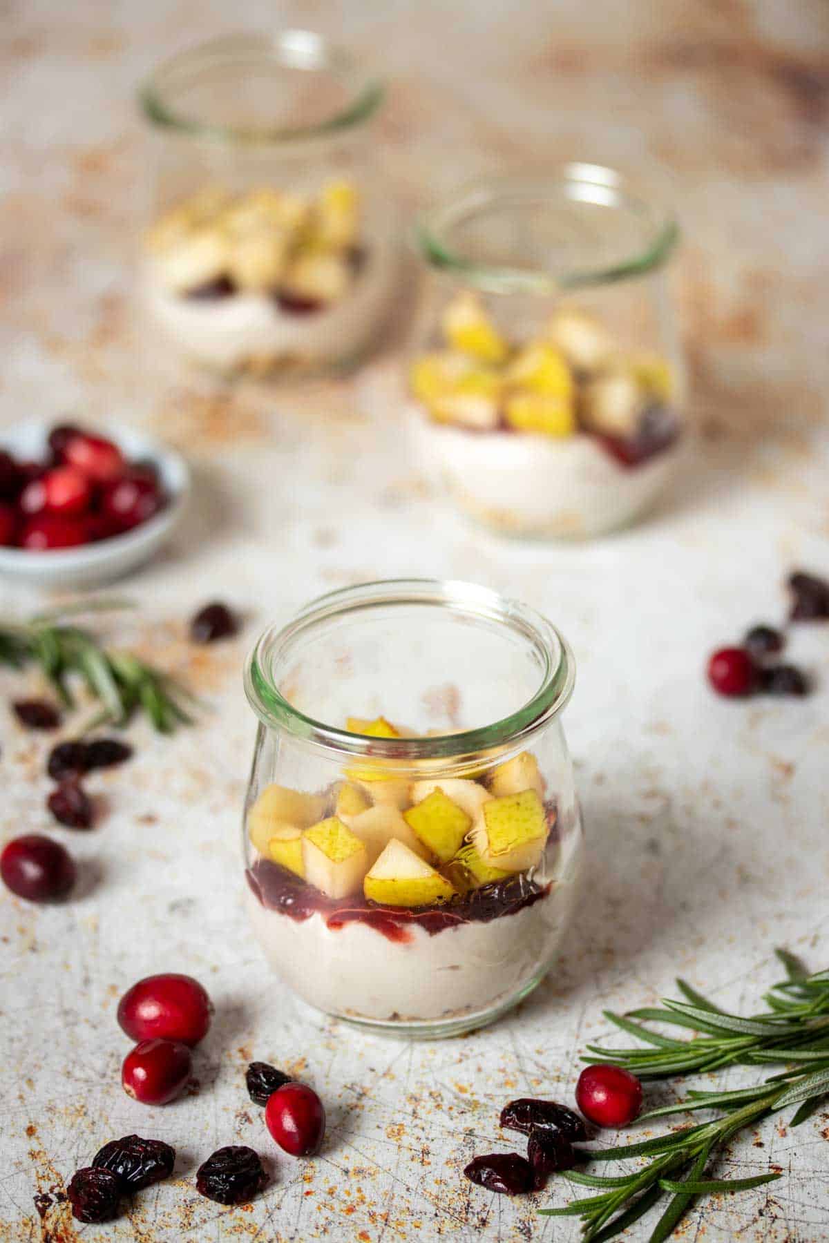 Yogurt, jelly and fruit layered in a glass jar in front of two other jars and cranberries and rosemary sprigs laying next to it.