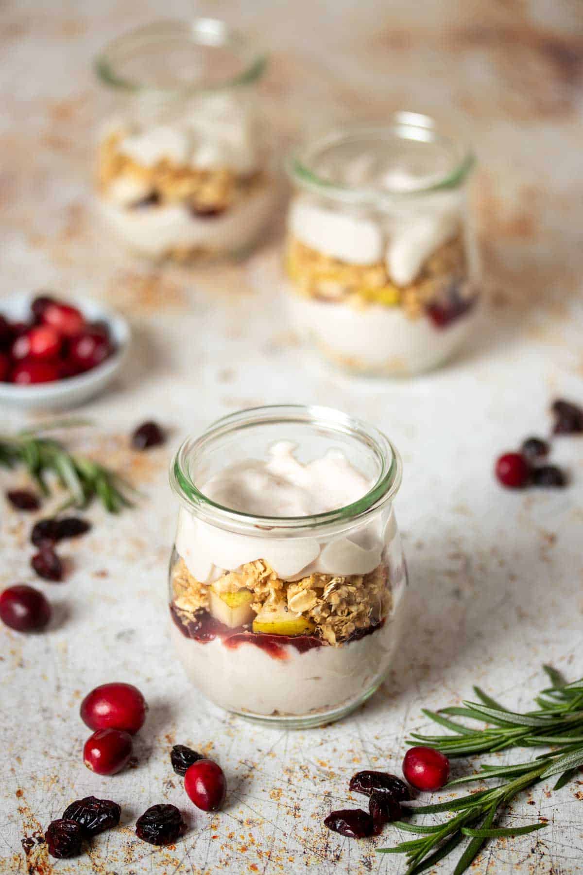 A glass jar layered with yogurt, jelly and granola with two other jars behind it and cranberries and rosemary sprigs on the surface.