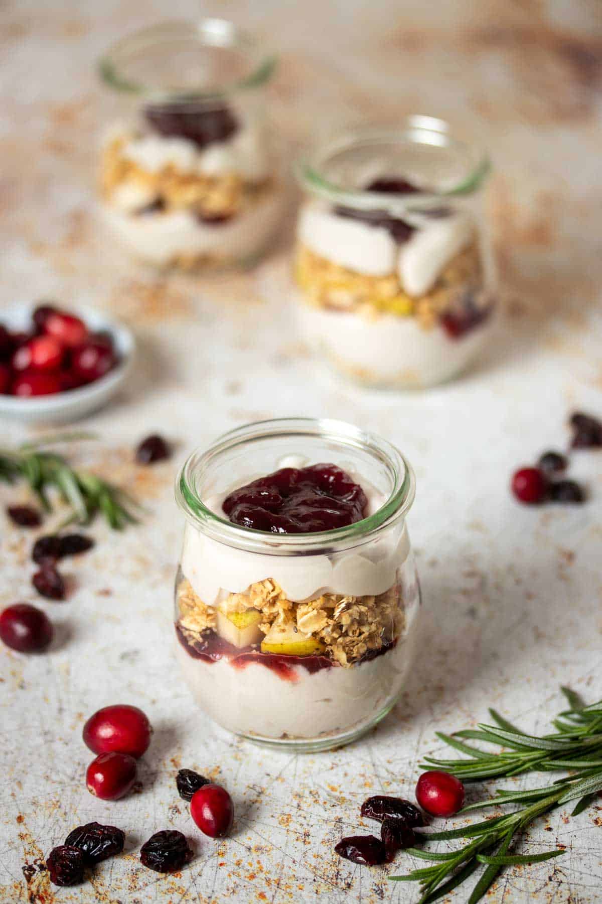 A glass jar layered with granola, yogurt, fruit and jelly and two more behind it.