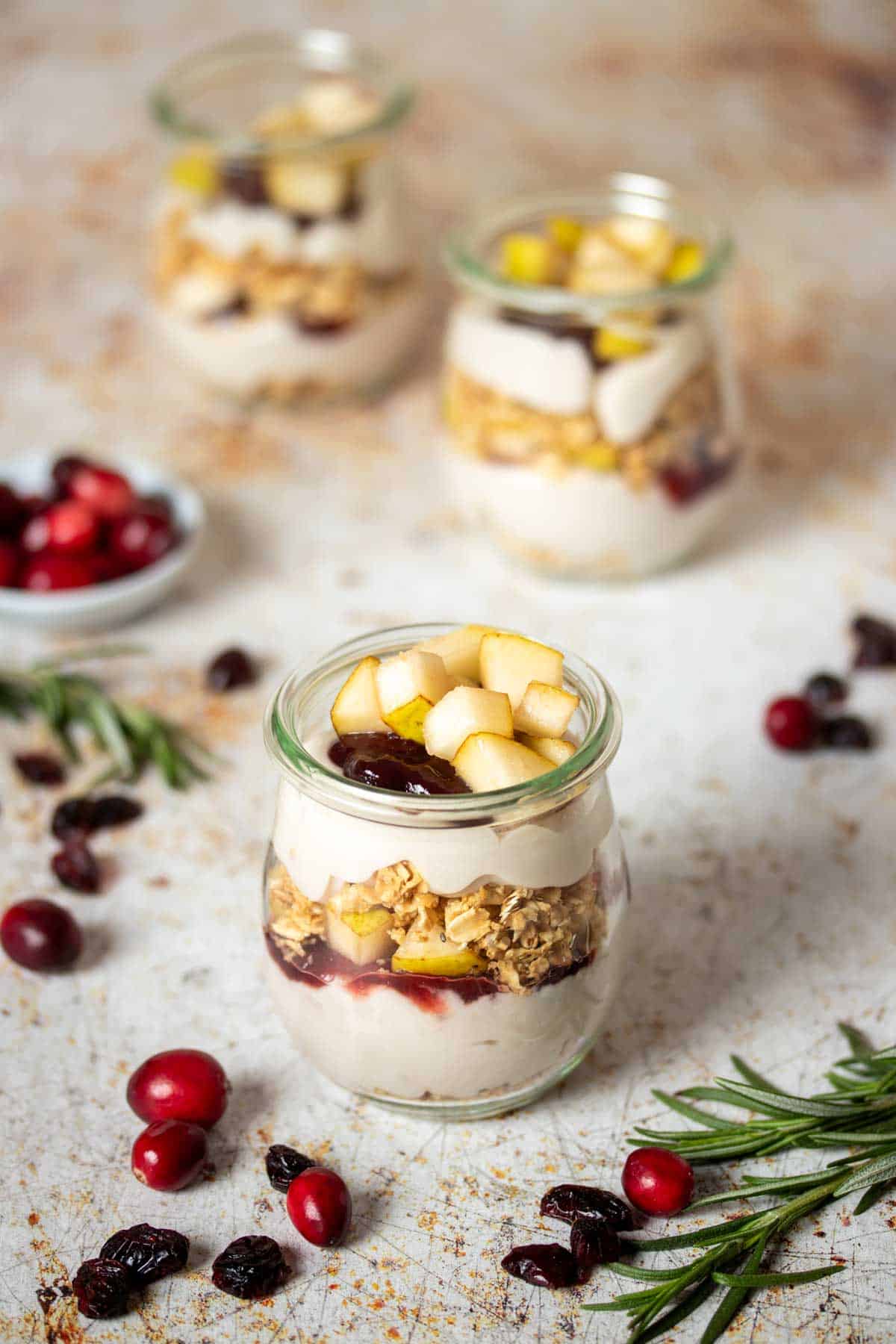 Three glass jars layered with yogurt, granola and fruit in two layers on a tan speckled surface.