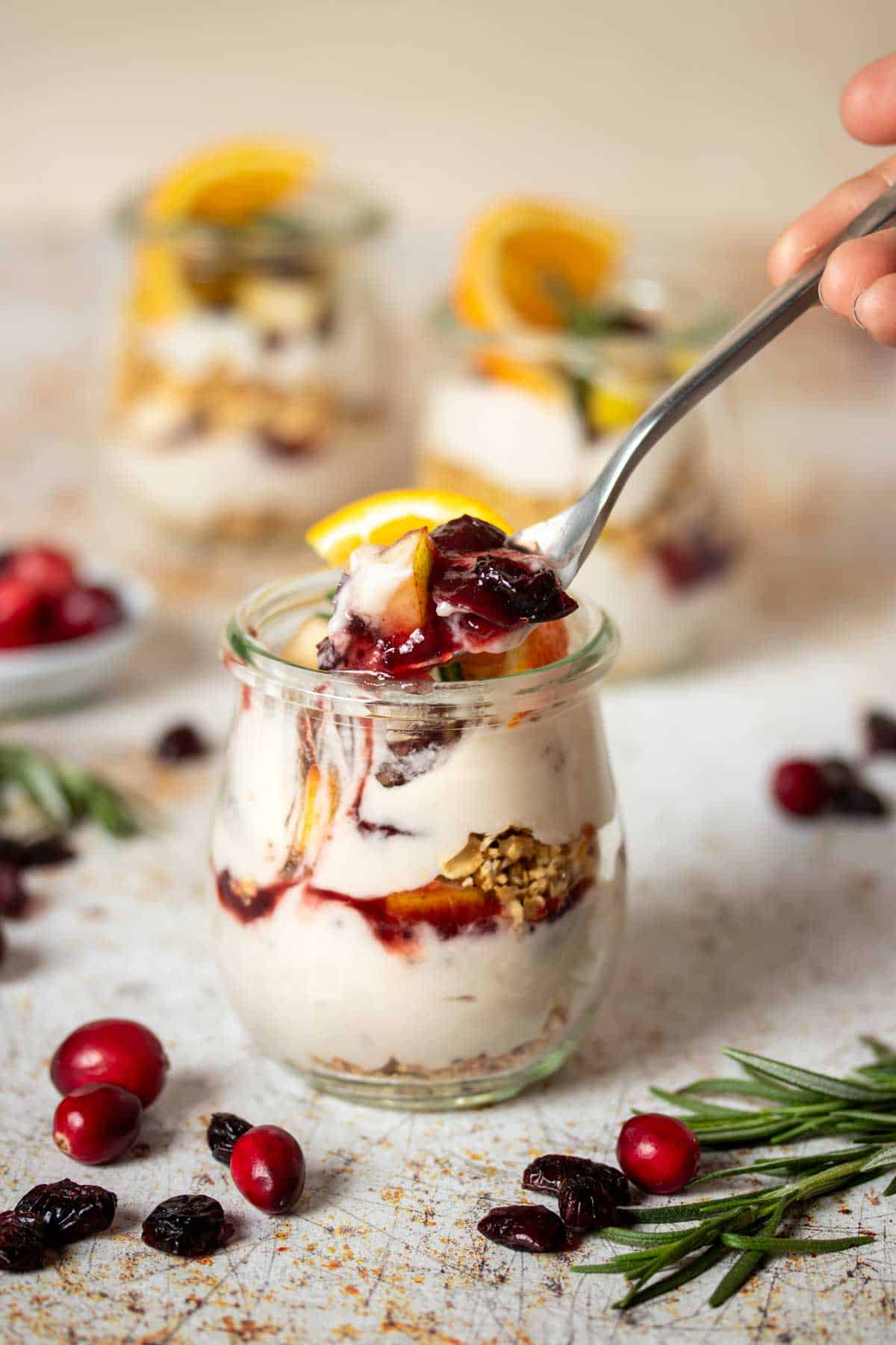 A spoon getting a bite from a glass jar with a yogurt granola fruit parfait in it in front of two more jars.