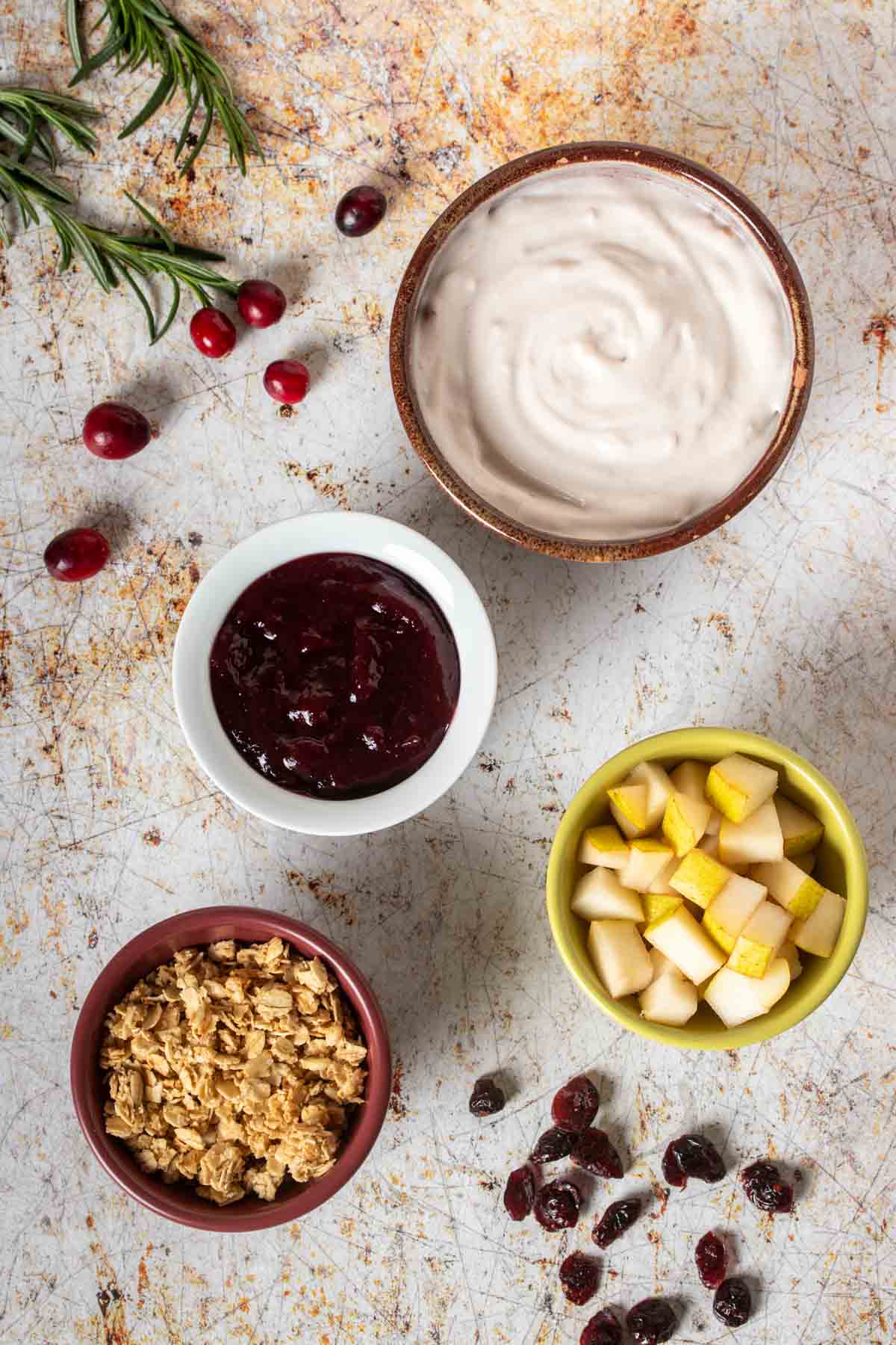 Different colors bowls on a tan speckled surface with jelly, yogurt, granola and chopped pears in them.