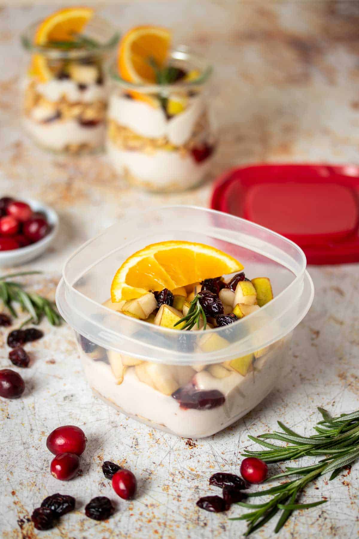 An open plastic container with layered yogurt granola parfait topped with fruit and two jars with the same filling behind it.