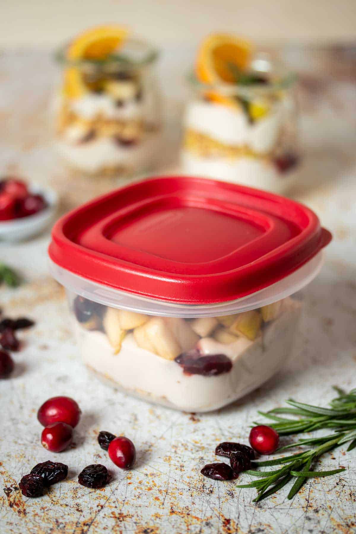 A plastic square container with a red top filled with layers of yogurt, granola and fruit in front of two filled jars and decor on the surface.