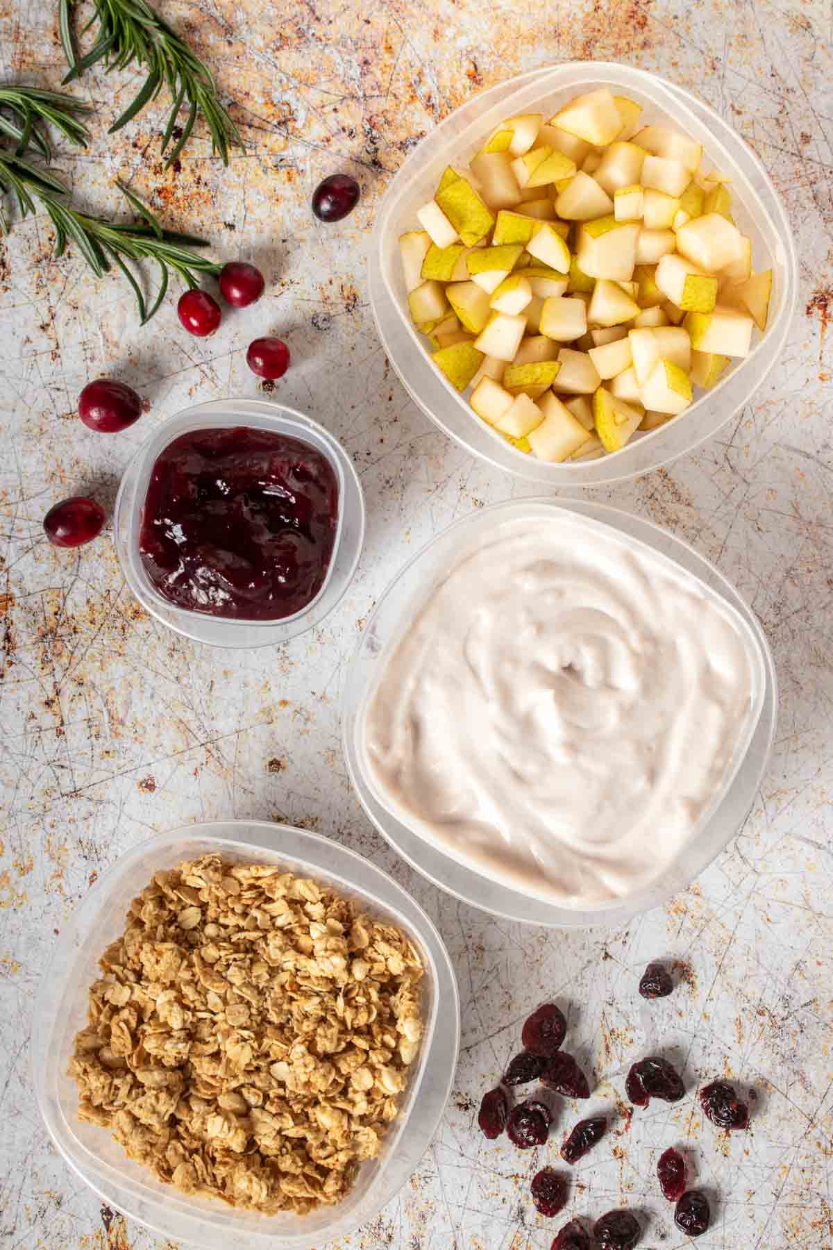 Top view of plastic containers filled with chopped pears, jelly, yogurt and granola sitting on a tan speckled surface.