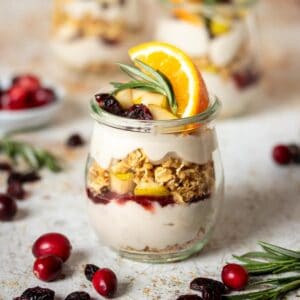 A fruit, yogurt and granola parfait in a glass jar topped with an orange slice and piece of rosemary on a speckled white brown surface with cranberries and rosemary sprigs next to it.