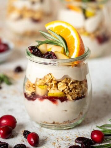 A fruit, yogurt and granola parfait in a glass jar topped with an orange slice and piece of rosemary on a speckled white brown surface with cranberries and rosemary sprigs next to it.