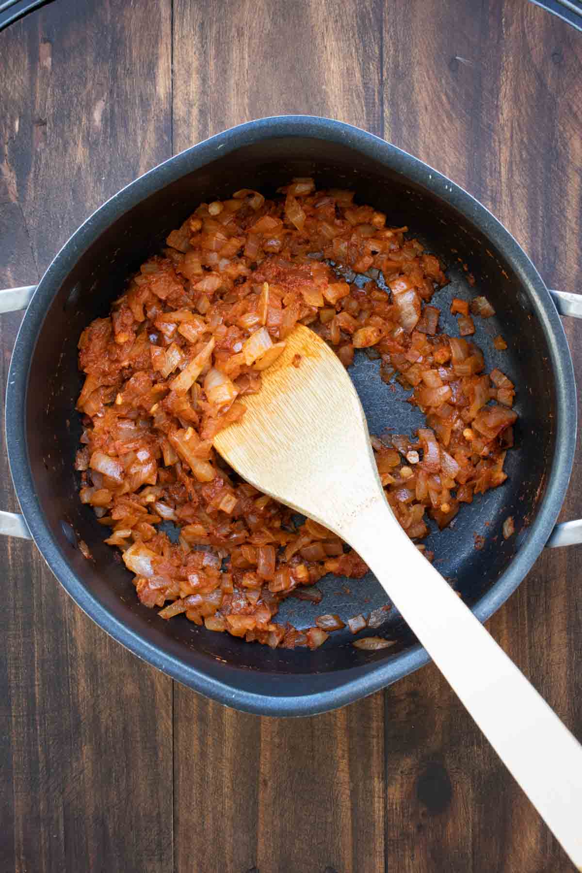 Wooden spoon stirring chopped onions and curry paste in a pot.