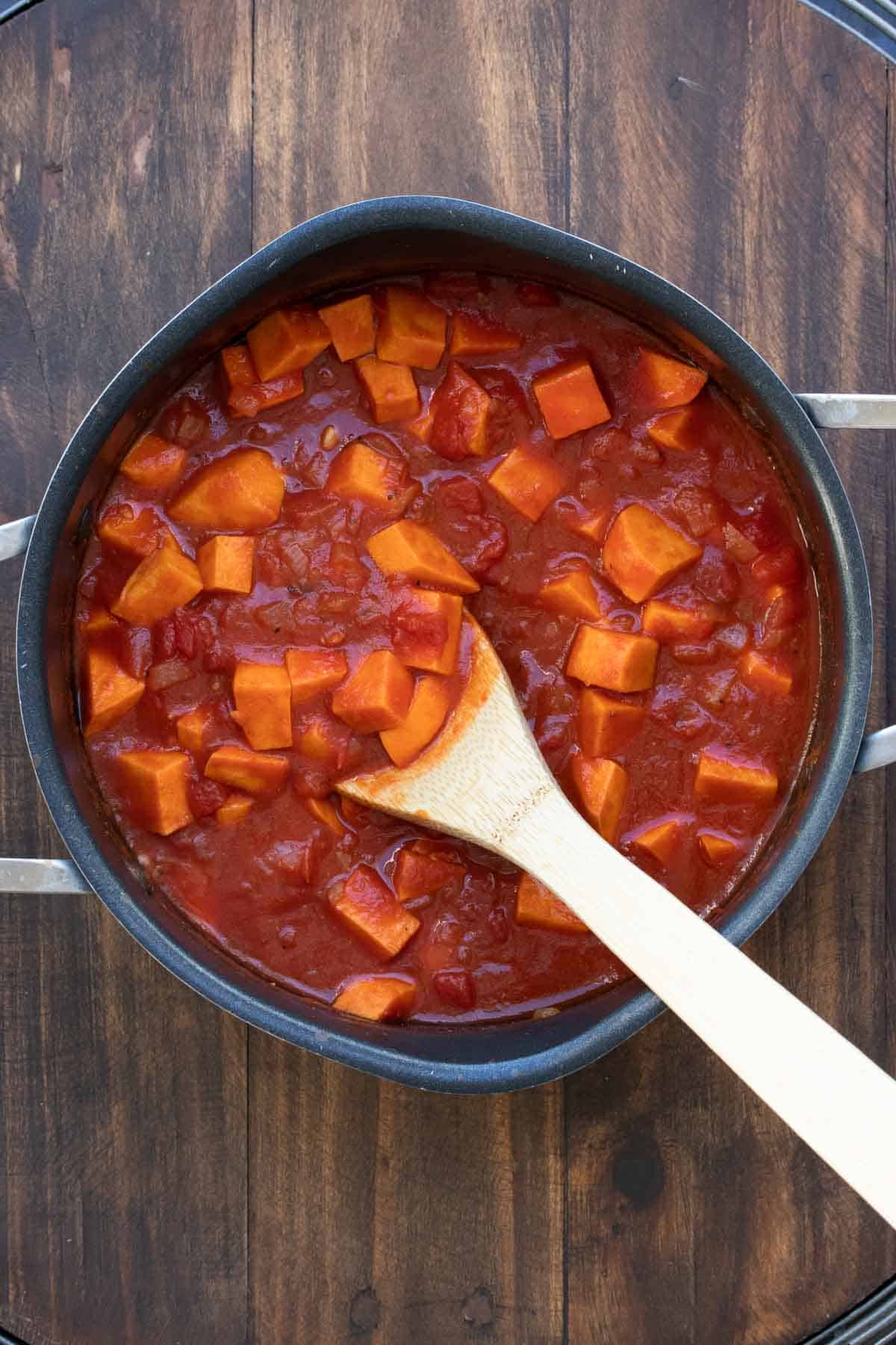 Chopped sweet potatoes in tomato broth being stirred in a pot.