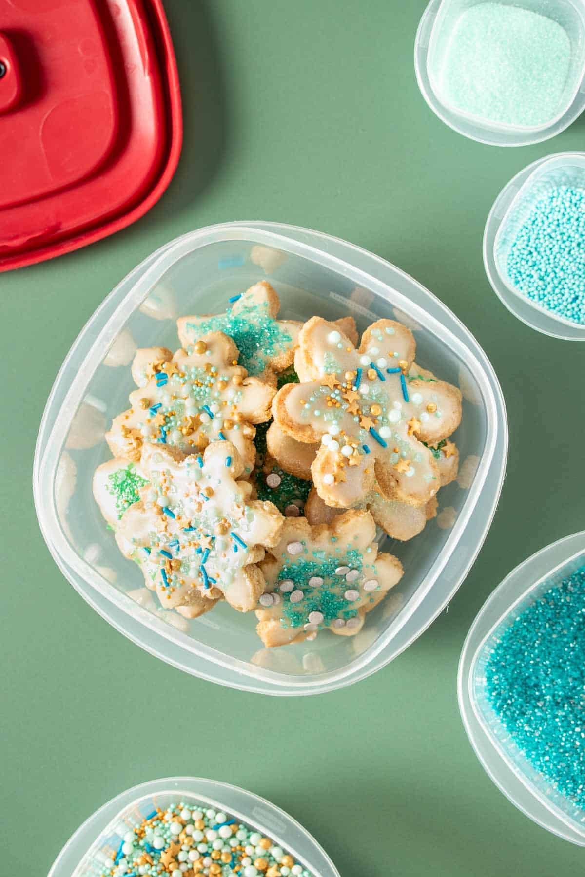 Top view of holiday sugar cookies decorated with green and blue sprinkles inside a plastic container with a red lid next to it.