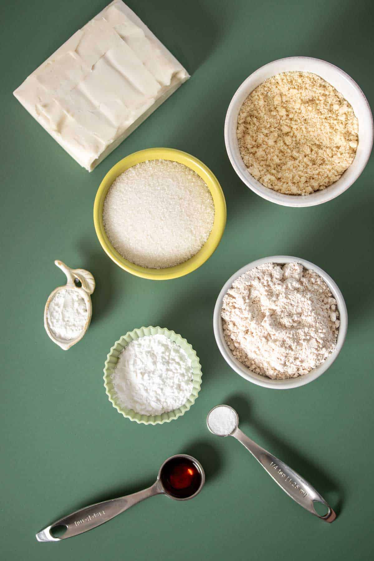 Different bowls with flours and sugar, measuring spoons with salt, baking powder and vanilla extract and a square of butter on a green surface.