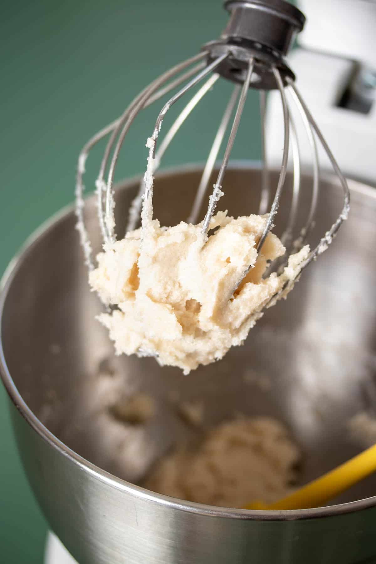 A stand mixer with the whisk attachment and a fluffy white butter sugar mixture on it.