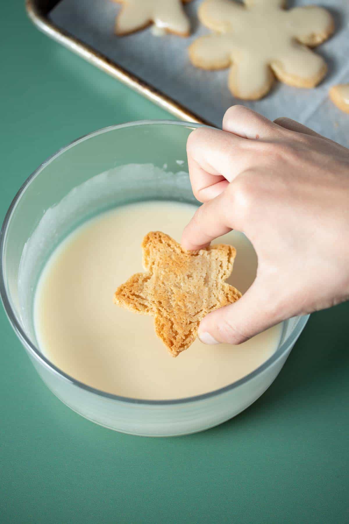 A hand dipping a star shaped sugar cookie in a glass bowl of white icing.