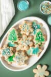 Sugar cookies in holiday shapes with green and blue decor on a white plate sitting on a green surface.