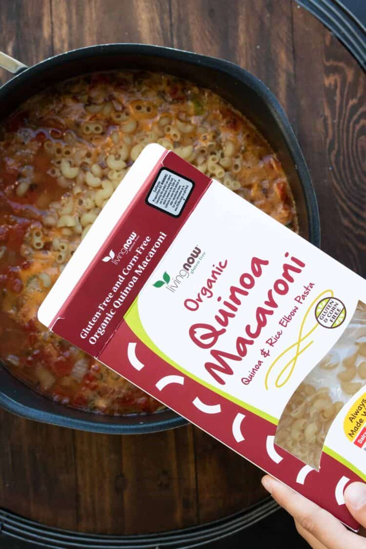 Hand pouring box of quinoa macaroni into a pot of soup.