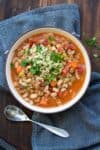 A white bowl with a pasta, bean and vegetable soup inside with a spoon next to it on a blue towel.