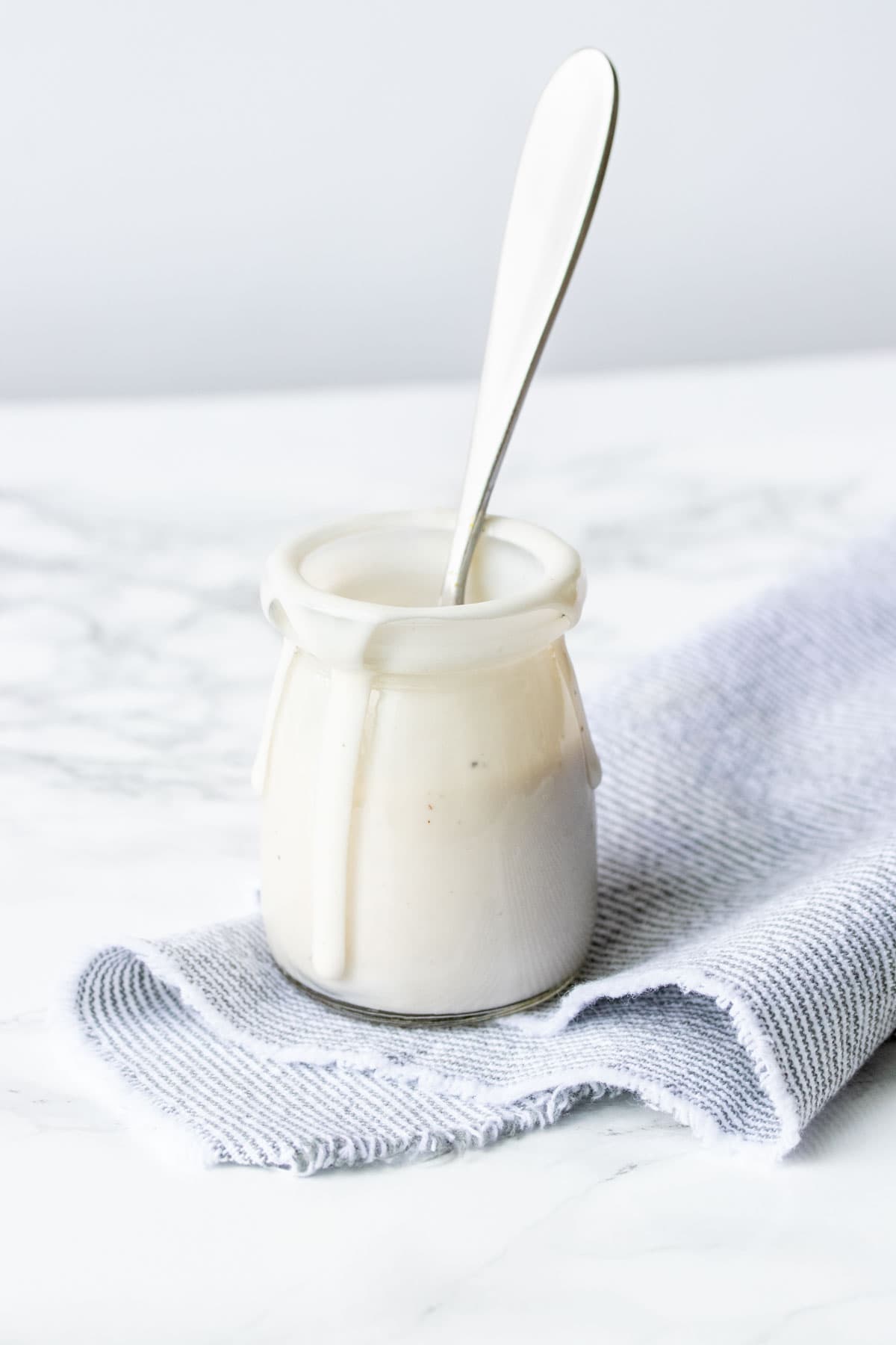 A glass jar with creamy sauce in it and a spoon on a marble grey surface.