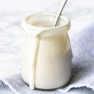 A close up of a glass jar with cashew cream sauce in it and dripping down the sides and a spoon inside.