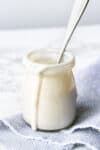 A glass jar with a spoon in it sitting on a grey striped towel filled with cashew cream sauce.