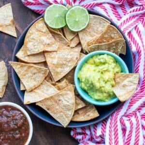 A black plate full of tortilla chips and a small bowl of guacamole sitting on a wooden surface with a red striped towel.