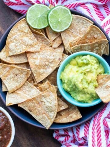 A black plate full of tortilla chips and a small bowl of guacamole sitting on a wooden surface with a red striped towel.